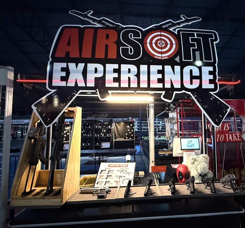 Front view of the AIRSOFT EXPERIENCE counter under its large sign, with a wooden rifle stand and pistols on display
