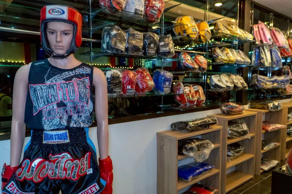 Mannequin in headgear, a singlet and black Muay Thai shorts with a BOXING STADIUM PATONG BEACH waistband, in front of the glove wall
