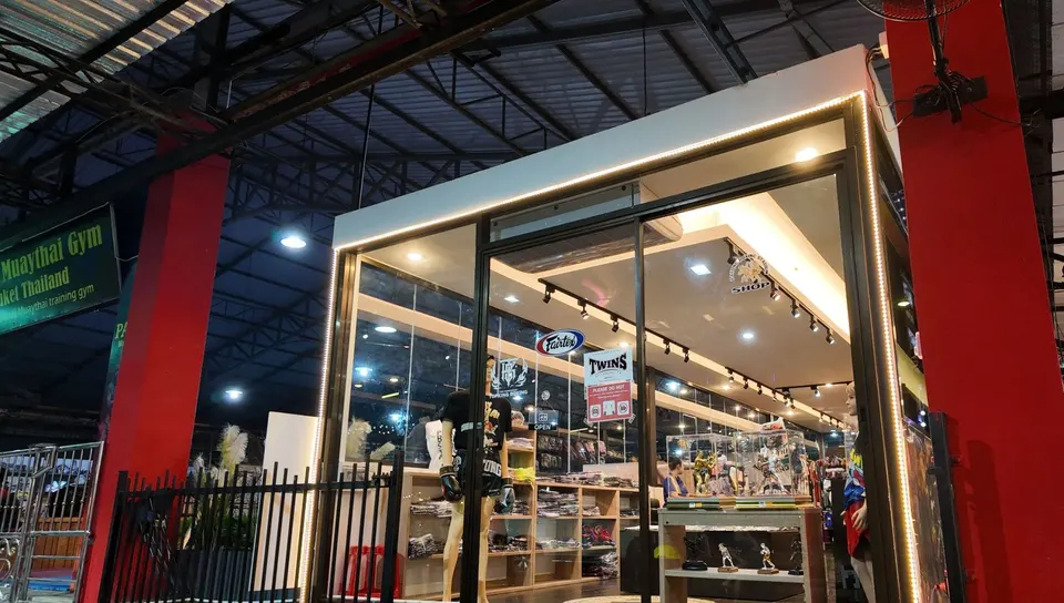The glass-fronted shop inside the stadium hall at night, with Fairtex and TWINS stickers on the door
