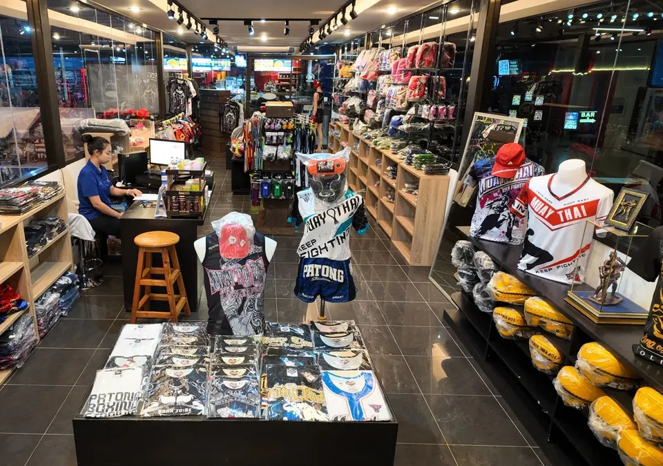 Inside the Muay Thai gear shop at the stadium: T-shirts on a display table, mannequins in stadium-branded shorts and shelves of gloves and pads